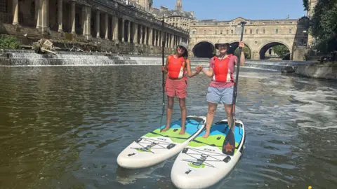 Alfie/Original Wild Two women are standing on paddleboards side by side on the water, posing for the camera and holding each other's hands. Bath's Pulteney Weir and Pulteney Bridge are behind them.