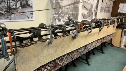 A large piece of machinery which used to stitch carpets is on display in a museum with black and white photos behind showing the machine in action.