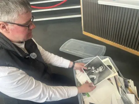 Brendan Bugg, a man with grey hair wearing glasses, a black gilet and a white shirt, kneels on a grey carpet and looks down at a box full to overflowing with publicity photos. He is holding a black and white image, probably dating from the late 1950s, of a man posing deliberately in a flat cap and tweed jacket.
