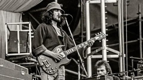 Getty A black and white image of a man performing on stage holding a Gibson SG guitar. He has long hair and is wearing a bucket hat.