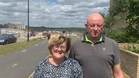 BBC/Mick Lunney A couple, Janice and Bob Bolan, pose for a photograph, the park site is in the background.