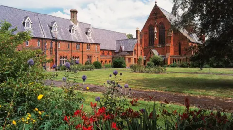 Royal Berkshire Archives A large three storey building made of red brick with a grey roof stands next to a church building. Before them is a green lawn with flowers and shrubs to the forefront of the image.
