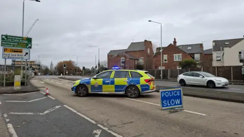 A police road block with a car parked across the approach to the bridge and a blue Police Slow sign set up