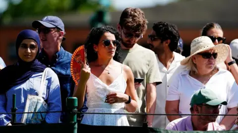 Spectators stand at Wimbledon - with one woman with curly dark hair and a white top standing in the middle holding a fan to cool down from the heat.