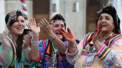 Three women clap, wearing colourful clothing and jewelry