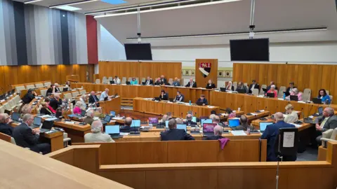 Paul Moseley/BBC We can see a mostly full Norfolk County Council chamber. It is a modern building but with varnished wood panels.
