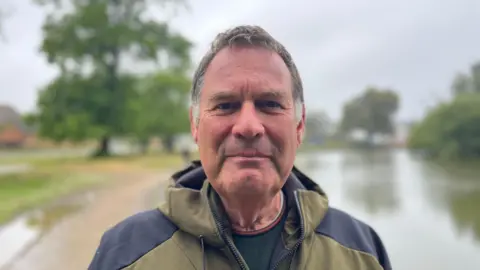 George Carden/BBC Russ Middleton who has brown hair wearing a green and black rain coat standing by Common Pond in hailsham 