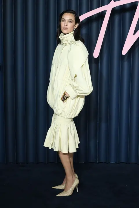 Getty Alexa Chung in a high-necked, long-sleeve cream dress that bunches on the thighs and flares over the knees. She wears cream-and-gold shoes.