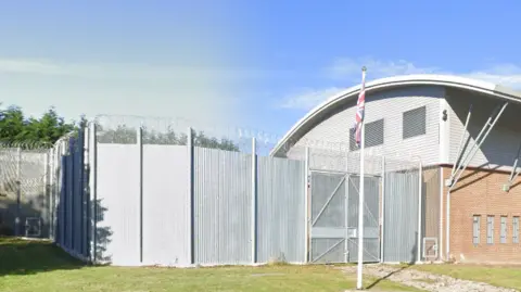 A view of the outside of HMP Deerbolt, a large fence topped with barbed wire can be seen next to a large building with a curved roof. A British flag can be seen on a flagpole outside.