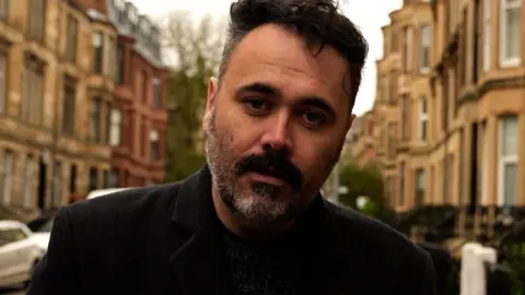 A man with short dark hair and a greying beard standing in a Glasgow tenement street.