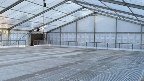 The inside of the winter ice rink which has been recently assembled. It shows a large white marquee with metal piping on the floor, before the structure has been flooded and frozen.