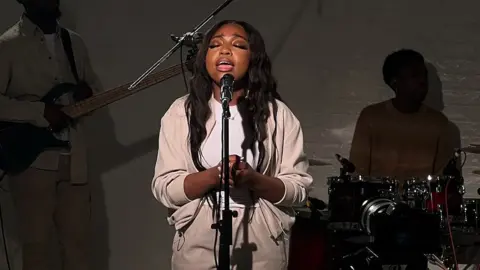 KAC Music Group Singer Chantelle Rutendo stood on stage singing into a black microphone. She is a black woman who her hands clasped together and has long black hair. There's a spotlight on her face and she has her eyes closed and her mouth open as she sings. She is wearing a white t-shirt and cream trousers and a cream open jacket. Behind her there's a silhouette visible of someone playing a guitar and someone playing the drums.