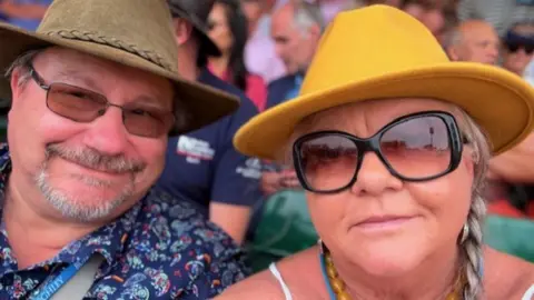Caroline Heaven Caroline Heaven, wearing sunglasses and a yellow hat, with her smiling husband Wayne, also wearing a hat and brightly coloured blue shirt, at a horse event