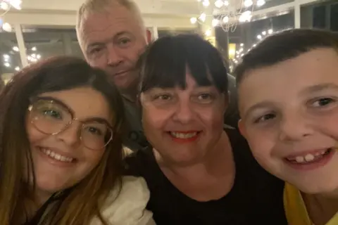 Ayla Jones Selfie photograph of Ayla (left) with her mother and father (centre) and her younger brother. The four of them all smile and look directly into the camera lens. Alya has brown hair, brown eyes and wears reading glasses. Her mother has a dark brown full fringe and brown eyes, while her father stands behind them in the background and smiles. He has silver short hair. 