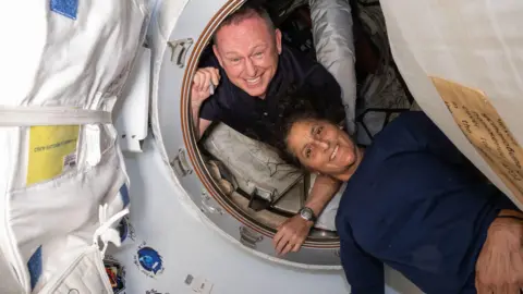 NASA Butch and Suni on the space station - both smiling down at the camera in a circular capsule and appearing to be in a suspended gravity environment. Both are dressed casually and the walls of the capsule appear to be made of a fabric-like material. 