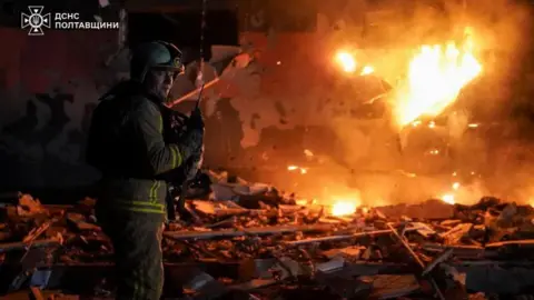 A firefighter works at the site of a building damaged during overnight Russian drone and missile strikes with flames seen in the background