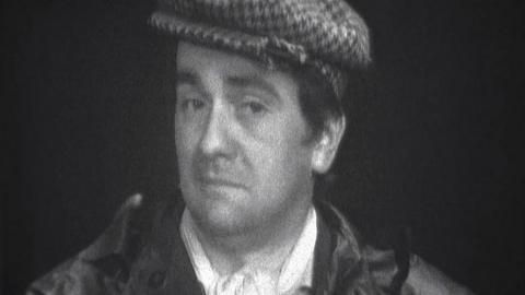 Dudley Moore in character as Dud, wearing a flat cap and looking unimpressed.
