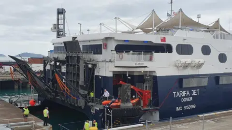 A blue and white DFDS ferry has arrived in the port. Staff in high-vis jackets stand nearby as the gangplank is lowered. 