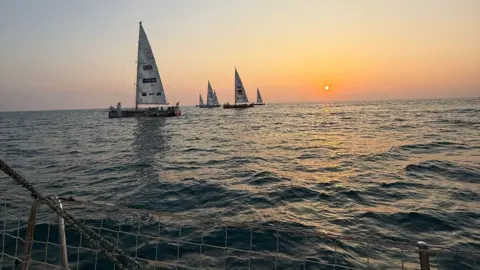 Bob Brown A sunset photo of boats in the sea. They have a white sail and the sun is low in the background. The sea looks calm. 