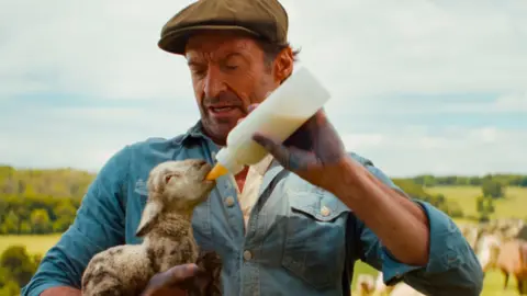 Hugh Jackman playing a shepherd. He's wearing a brown flat cap with a blue jacket with rolled up sleeves. Underneath he has a white shirt. He's looking down at and cradling a muddy lamb in his right arm while feeding it with a plastic milk bottle.