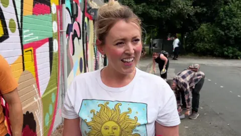 A woman wearing a white T-shirt with a yellow image of a sun on it. She has blonde hair, earrings and is standing in front of a building with colourful artwork.