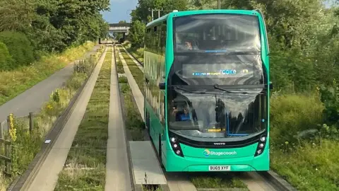 A double-decker bus on a guided busway heading towards the viewer along concrete tracks and kerbs in 2021. It has bright green livery. It is branded Stagecoach. It is travelling along a concrete track. On its right is a cycle and walkway. On either side are trees. 