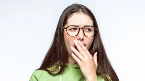 Getty Images Woman, with dark hair and brown glasses wearing a green top, is looking off to the side and has her hand up to her face in a yawn