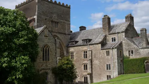 PA Media Buckland Abbey on a sunny day. It is a large grey bricked building with several levels and windows. Trees and a grass lawn are in front of the building.