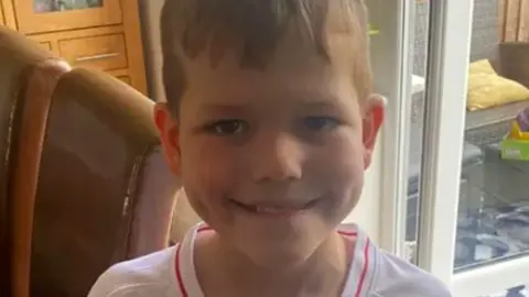 Eight-year-old Joshua Hillstead, grinning, and wearing a white t-shirt, while standing in front of some patio doors