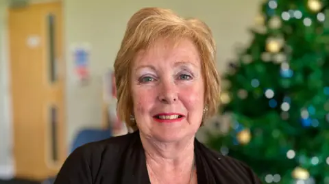 Judi Buck is smiling. She has short ginger hair and is wearing red lipstick and a black top. She in front of a Christmas tree which has blue lights and gold baubles on it.