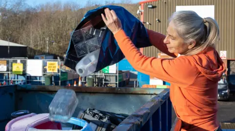 A woman with a silver/blonde ponytail and an orange hoodie. She is standing in front of a large metal recycling container. She is tipping a navy bag into it. Clear plastics are falling into the container.