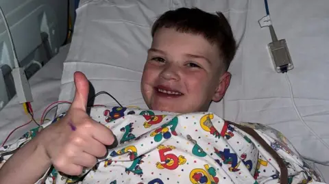 Dáithí is lying in a hospital bed with tubes around him. He has short brown hair, smiling at the camera and is doing the thumbs up - he is wearing a patterned hospital gown with coloured numbers and cartoons. 