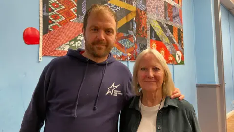 Alexa Rusakoff / BBC Tim Holtam and Liz Fletcher are standing side by side in front of a patterned fabric artwork on a blue wall. Holtam is wearing a dark hoodie with the club logo and Fletcher is wearing a light shirt and jacket.