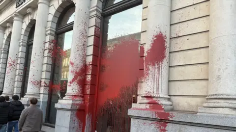 The outside of a bank building with large pillars either side of high windows. There is a red substance which looks like paint on the window and splashed on the walls.