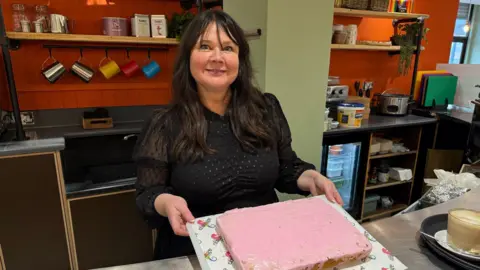 Sarah Farrell-Forster smiling into the camera as she holds a cake decorated with pink icing. She has long black hair and is wearing a black dress.