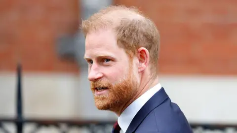 Prince Harry, a man with ginger hair and a bear, wearing a navy suit jacket with a white shirt and a tie. He is looking to the side and the background behind him is blurred. 