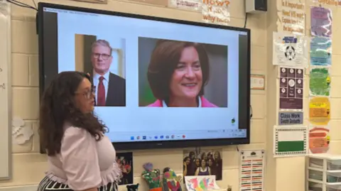 Woman stands next to a classroom projector with images of Keir Starmer and Eluned Morgan
