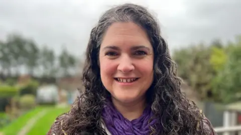 A woman wtih dark curly hair standing in from of her garden on a grey, drizzly day. She's smiling at the camera and wearing a purple crocheted scarf. To her right a garden path runs down towards outbuildings and tall trees which are blurry in the background. 