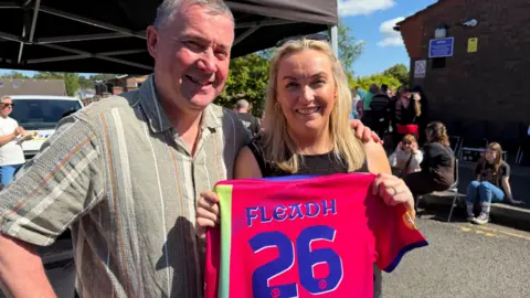 A man, with short grey hair, wearing a short sleeve green shirt, with his hand around a woman, with long blonde hair. The woman is holding up a pink jersey with 'Fleadh 26' written on the back. 