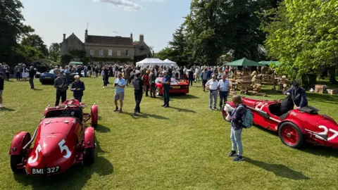 An image showing the event Middlewick House. There are multiple red classic cars on display with a crowd of people on the field. There are also white and green stalls up