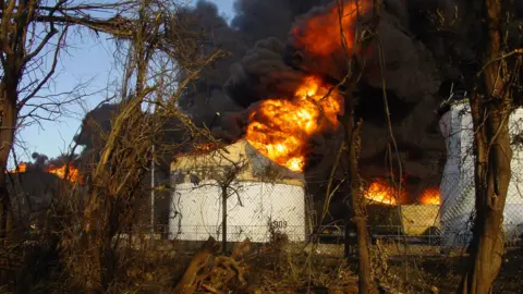 David Humphreys Large industrial fire with orange flames and black smoke engulfing white storage tanks behind a chain-link fence. Surrounding trees and bushes are brown and dries out,