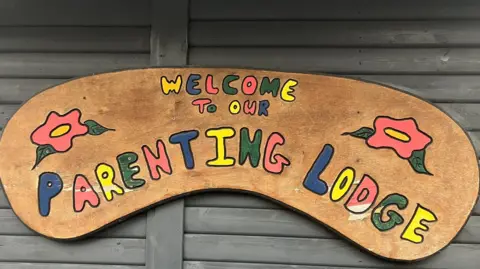 A handmade sign which reads 'Welcome to our Parenting Lodge' in colourful writing with flowers either side is attached to a wall