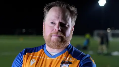 Jamie Niblock/BBC Peter Jenkyn looks seriously at the camera. He has reddish short hair and a beard, and is wearing a yellow and blue football jersey. He is standing on a football pitch.