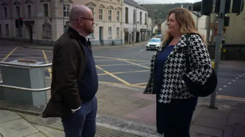 A man in a green coat, David Grundy, speaks to a woman wit blonde hair and a white and black coat, Sioned Williams, in a town centre