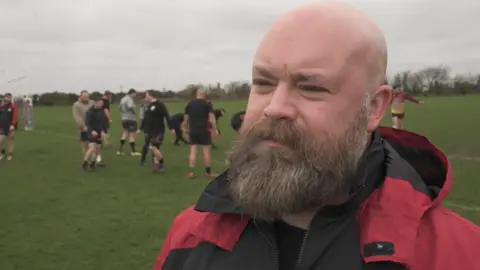 Dean is being interviewed next to the rugby pitch, with a group of players seen training behind him. He is wearing a red windbreaker jacket, and has a bald head and a beard.