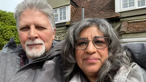 Maria Kelly A man with grey hair and a beard in a grey jacket next to a woman with dark hair and glasses in a grey jacket with a house behind them