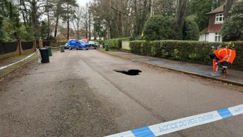 Nicola Haseler/BBC A residential street is cordoned off with blue-and-white police tape. A large hole or sinkhole is visible in the middle of the road.