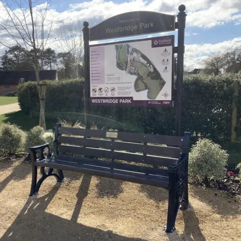 LDRS A memorial park bench in a park placed in front of a hedge and plants. A large wooden Westbridge Park sign is behind the bench featuring a map of the park.