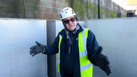 David Nuth wearing a white hard hat, a bright yellow safety vest, dark gloves, and a navy jacket standing outdoors in an industrial harbour setting. He's standing between large pale-coloured panels and extending both arms outward in an open gesture. Behind him is a tall stone wall with green staining from exposure to water and weather.
