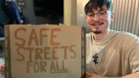 Jon Wright/BBC A man sits inside a cafe holding up a sign which reads Safe streets for all. All is underlined.